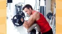 fit, focused man in red tank top and black gym shorts lifting barbell in bright gym