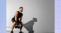 muscular man lifting two kettlebells outdoors in front of cement wall