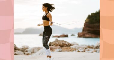 fit brunette woman in black sports bra and black and white leggings doing jump rope workout at the beach