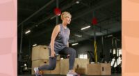 happy mature woman in gray tank top and dark gray leggings doing dumbbell lunges at the gym