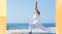 fit middle-aged woman in breezy white yoga clothes doing crescent lunge on mat by the beach