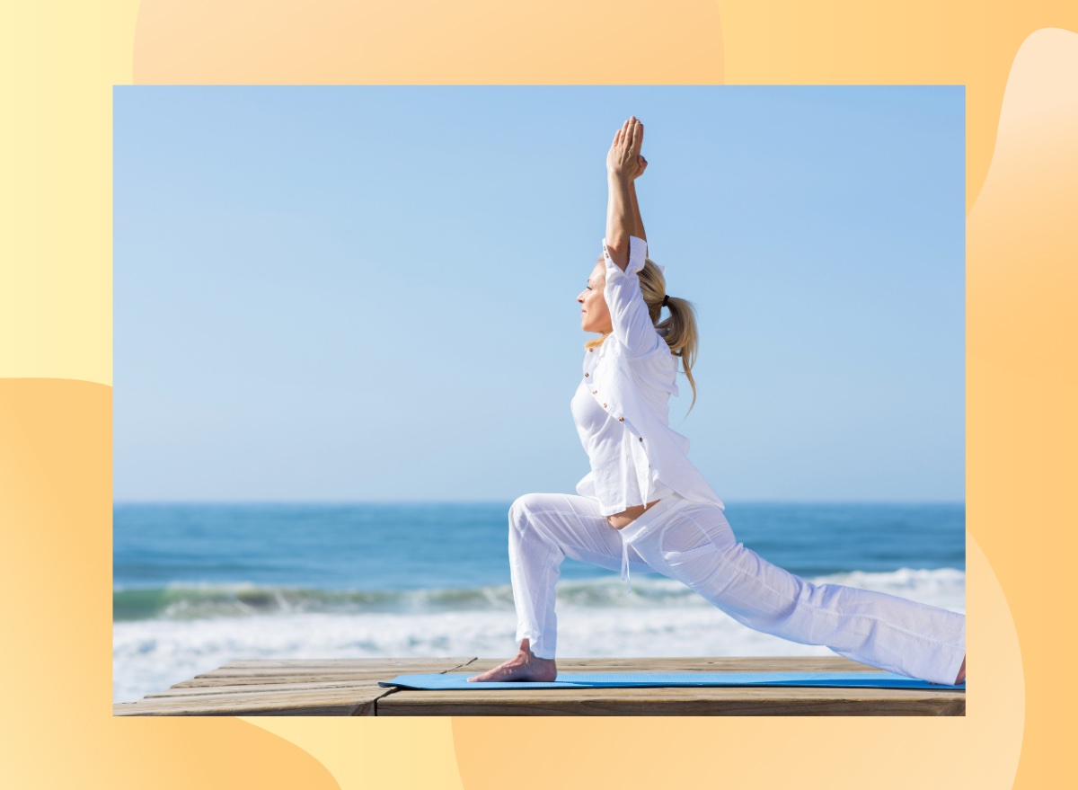 fit middle-aged woman in breezy white yoga clothes doing crescent lunge on mat by the beach