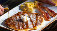 person pouring syrup on first watch pancake with a side of eggs and bacon