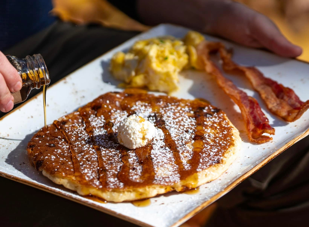 person pouring syrup on first watch pancake with a side of eggs and bacon