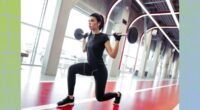 focused brunette woman doing barbell lunge on an indoor track at the gym