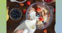 close-up of hand making fresh fruit protein shake with milk, chocolate protein powder, and berries