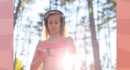 blonde woman wearing headphones and pink sports bra checking fitness tracker while on a run through the woods on sunny day