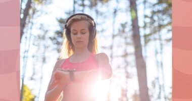 blonde woman wearing headphones and pink sports bra checking fitness tracker while on a run through the woods on sunny day