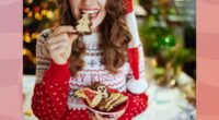 close-up of brunette woman in holiday sweater and Santa hat eating a Christmas tree cookie and holding plate of cookies