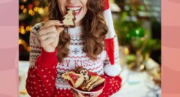 close-up of brunette woman in holiday sweater and Santa hat eating a Christmas tree cookie and holding plate of cookies