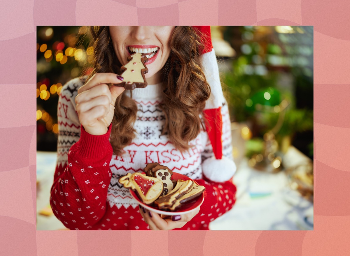 close-up of brunette woman in holiday sweater and Santa hat eating a Christmas tree cookie and holding plate of cookies