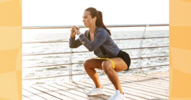 fit brunette woman doing resistance band squat exercise on boardwalk by the ocean