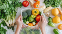 Close-up of hands placing fresh vegetables and fruits into an eco-mesh bag on a white table.