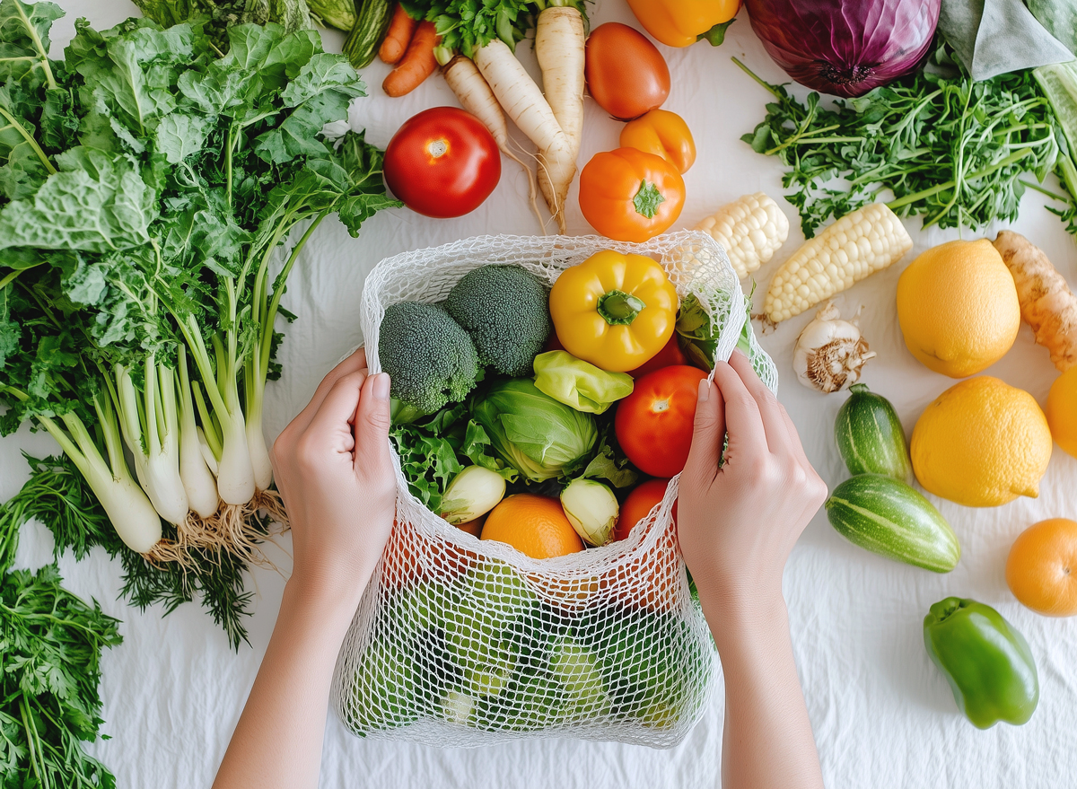 Close-up of hands placing fresh vegetables and fruits into an eco-mesh bag on a white table.