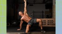 fit, muscular, shirtless man doing Turkish get-up exercise in dark gym set-up