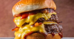 double meet Burger with melting cheddar cheese and bacon centralized on a gray surface and brown background.