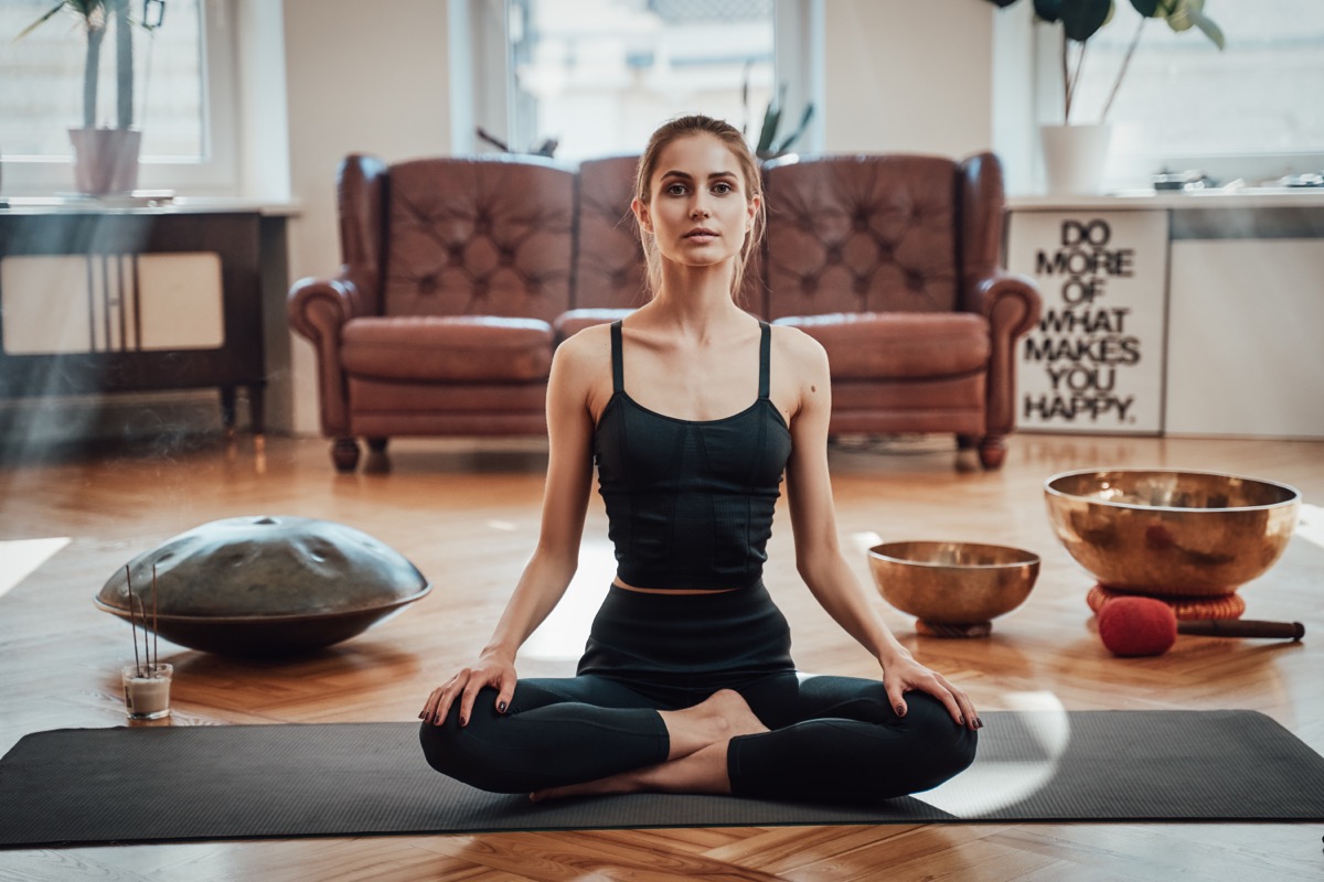 Relaxed caucasian woman dressed in black sportswear sits on carpet in lotos pose looking at camera in shiny and atmospheric living room.