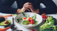 Weight loss plans for women theme white ceramic bowl contained healthy salad (slided avocados, boiled eggs, rocket leaves, tomatoes) on wooden table with blurred woman in black dress / selective focus