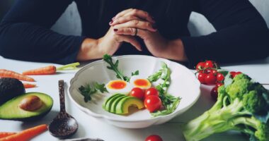 Weight loss plans for women theme white ceramic bowl contained healthy salad (slided avocados, boiled eggs, rocket leaves, tomatoes) on wooden table with blurred woman in black dress / selective focus