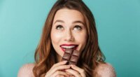 Portrait of a delighted brown haired woman with bright makeup eating chocolate bar