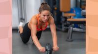 fit brunette woman in orange sports bra and black workout leggings doing an ab wheel rollout at the gym