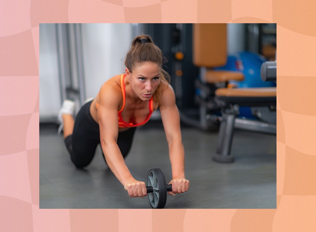 fit brunette woman in orange sports bra and black workout leggings doing an ab wheel rollout at the gym