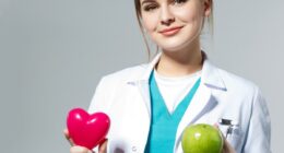 Beautiful smiling female doctor holding red heart and green apple in front of chest. Health life and wholesome food concept. Vegetarian lifestyle concept. Cardiology concept