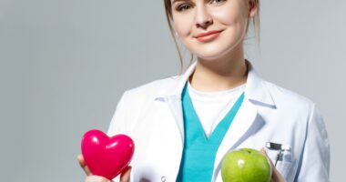 Beautiful smiling female doctor holding red heart and green apple in front of chest. Health life and wholesome food concept. Vegetarian lifestyle concept. Cardiology concept