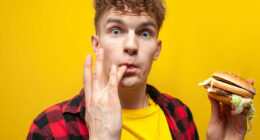 young surprised guy student eats a big tasty burger on a yellow background and licks the sauce on his finger, a man eats fast food and looks at the camera