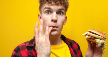 young surprised guy student eats a big tasty burger on a yellow background and licks the sauce on his finger, a man eats fast food and looks at the camera