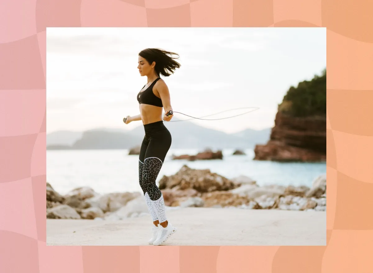 fit brunette woman in black sports bra and black and white leggings doing jump rope workout at the beach